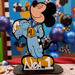 May include: A Mickey Mouse-themed cake topper. The cartoon figure is dressed in a blue racing suit and holds a helmet. A gold medal with the number 1 is around his neck. The base has the name "Noa" and the number 1 in white.