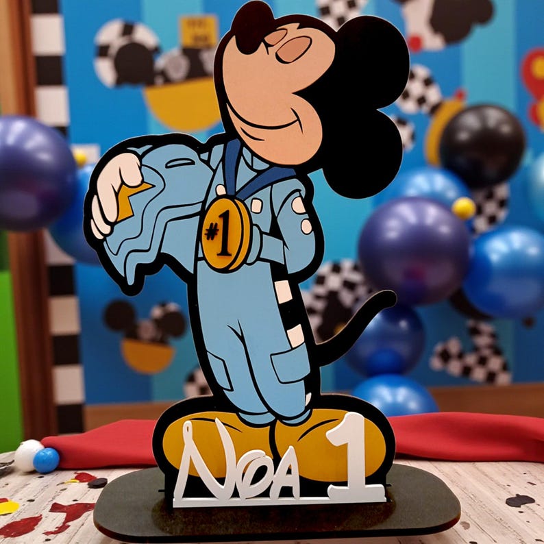 May include: A Mickey Mouse-themed cake topper. The cartoon figure is dressed in a blue racing suit and holds a helmet. A gold medal with the number 1 is around his neck. The base has the name "Noa" and the number 1 in white.