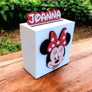 Op de afbeelding: Een witte houten doos met een gleuf aan de bovenkant, met een Minnie Mouse-ontwerp en de naam "Joanna" in rood en wit. De doos heeft een Minnie Mouse-gezicht met een rode strik met stippen.