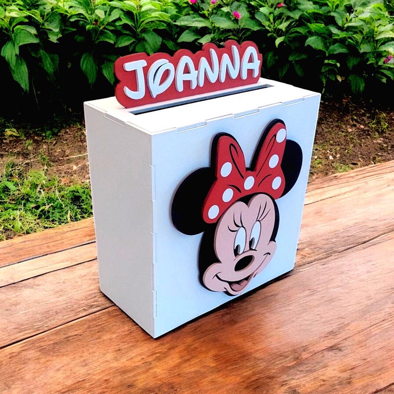 Op de afbeelding: Een witte houten doos met een gleuf aan de bovenkant, met een Minnie Mouse-ontwerp en de naam "Joanna" in rood en wit. De doos heeft een Minnie Mouse-gezicht met een rode strik met stippen.