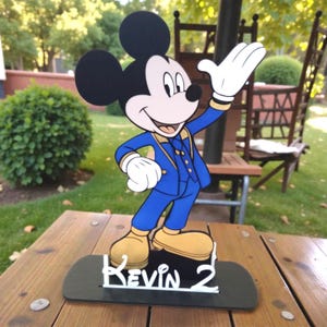 K&ouml;nnte beinhalten: Ein Holzausschnitt von Mickey Mouse in einem blauen Anzug mit goldenen Akzenten, der auf einer schwarzen Basis steht. Die Basis tr&auml;gt den Namen "Kevin 2" in wei&szlig;er Schrift. Der Hintergrund ist eine verschwommene Au&szlig;enszene.
