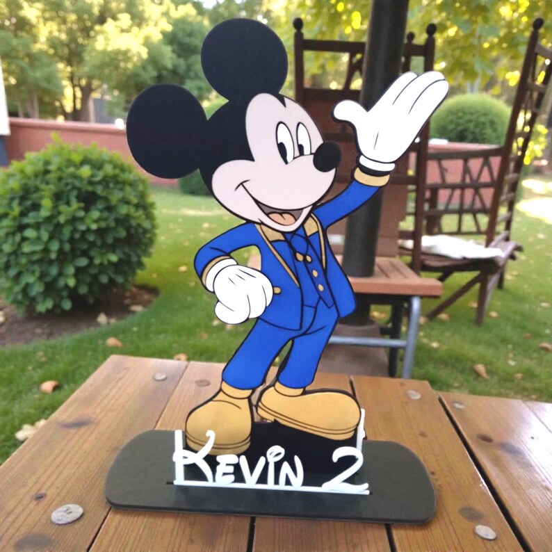 K&ouml;nnte beinhalten: Ein Holzausschnitt von Mickey Mouse in einem blauen Anzug mit goldenen Akzenten, der auf einer schwarzen Basis steht. Die Basis tr&auml;gt den Namen "Kevin 2" in wei&szlig;er Schrift. Der Hintergrund ist eine verschwommene Au&szlig;enszene.