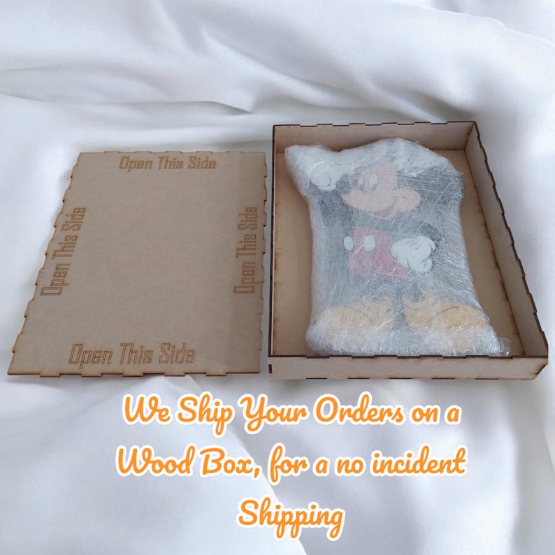 K&ouml;nnte beinhalten: Eine Holzkiste mit einem lasergeschnittenen Deckel, der mit "Open This Side" beschriftet ist. Im Inneren ist eine verpackte Mickey Mouse-Figur zu sehen. Der Text auf dem Bild lautet "We Ship Your Orders on a Wood Box, for a no incident Shipping."