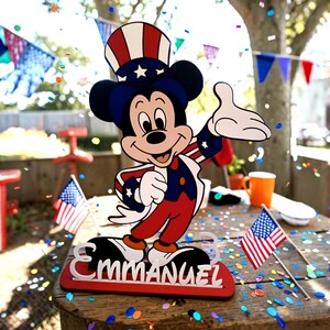 Puede incluir: Figura de madera de Mickey Mouse con atuendo patri&oacute;tico en rojo, blanco y azul. La figura est&aacute; sobre una base con el nombre "EMMANUEL" en letras blancas. Banderas americanas y confeti dispersos sobre una mesa de madera.