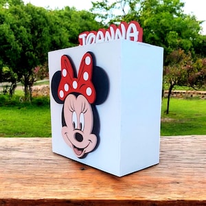 Op de afbeelding: Een witte doos met een Minnie Mouse-ontwerp. De doos heeft een rode strik met witte stippen en de naam "TOANNA" in het rood. De doos staat op een houten oppervlak.