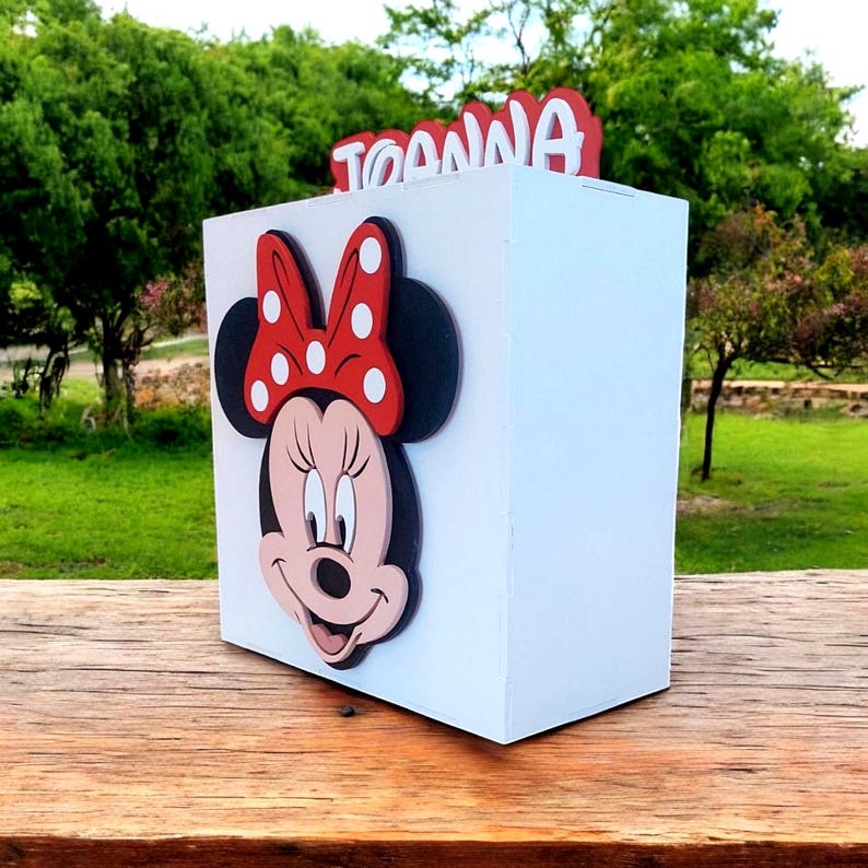 Op de afbeelding: Een witte doos met een Minnie Mouse-ontwerp. De doos heeft een rode strik met witte stippen en de naam "TOANNA" in het rood. De doos staat op een houten oppervlak.