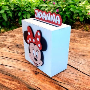 Op de afbeelding: Een witte houten doos met een gleuf aan de bovenkant, met een Minnie Mouse-ontwerp op de voorkant. De naam "JOANNA" wordt in rode letters boven de gleuf weergegeven. De doos staat op een houten oppervlak.