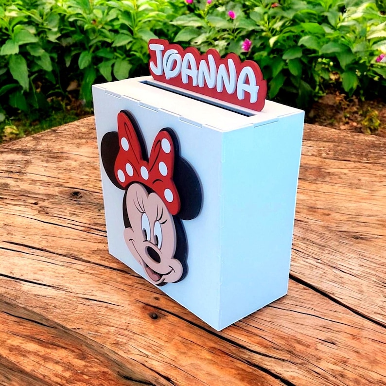 Op de afbeelding: Een witte houten doos met een gleuf aan de bovenkant, met een Minnie Mouse-ontwerp op de voorkant. De naam "JOANNA" wordt in rode letters boven de gleuf weergegeven. De doos staat op een houten oppervlak.