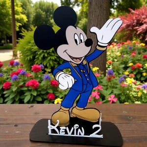 K&ouml;nnte beinhalten: Eine Mickey Mouse-Figur in einem blauen Anzug mit goldenen Akzenten, wei&szlig;en Handschuhen und beigefarbenen Schuhen. Die Figur steht auf einer schwarzen Basis mit dem Namen "Kevin 2" in wei&szlig;er Schrift. Der Hintergrund zeigt bunte Blumen und Gr&uuml;n.
