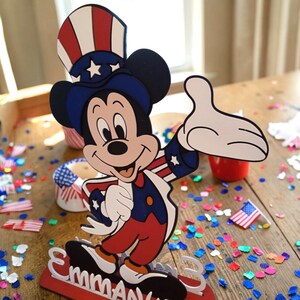 Puede incluir: Un recorte de Mickey Mouse en colores patri&oacute;ticos, con sombrero de copa y chaqueta, con el nombre "Emma" en la base. La escena incluye confeti y peque&ntilde;as banderas americanas, creando un ambiente festivo.