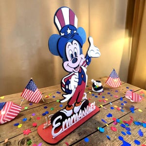 Puede incluir: Una figura patri&oacute;tica de Mickey Mouse en rojo, blanco y azul, con un gesto de pulgar hacia arriba. La figura se encuentra sobre una base roja con el nombre "Emmanuel" en blanco. Banderas americanas y confeti rodean la figura.