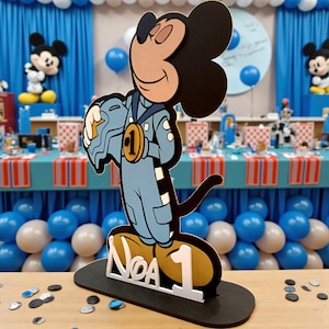 May include: A Mickey Mouse-themed birthday decoration. The cutout features Mickey in a racing suit, holding a helmet, and wearing a gold medal. The base reads "Noa 1". The color scheme includes blue, black, white, and gold.