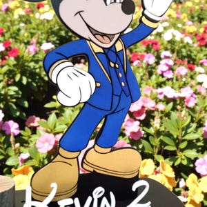 K&ouml;nnte beinhalten: Eine Mickey Mouse-Figur in einem blauen Anzug mit gelben Akzenten, die vor einem bunten Blumenbeet steht. Die Figur hat wei&szlig;e Handschuhe und hellbraune Schuhe. Der Name "Kevin" und die Zahl "2" sind unten sichtbar.