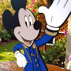 K&ouml;nnte beinhalten: Ein Holzausschnitt von Mickey Mouse in einer blau-goldenen Uniform, der mit einer wei&szlig;en behandschuhten Hand winkt. Die Figur hat schwarze Ohren, ein hellbraunes Gesicht und eine schwarze Nase. Der Hintergrund zeigt Gr&uuml;n und rosa Blumen.