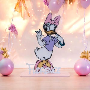 May include: A Daisy Duck-themed party decoration. The cartoon character is in a purple dress and pink bow. The name "FLORENCE" is displayed on a pink base. The background includes pink balloons and decorative lights.