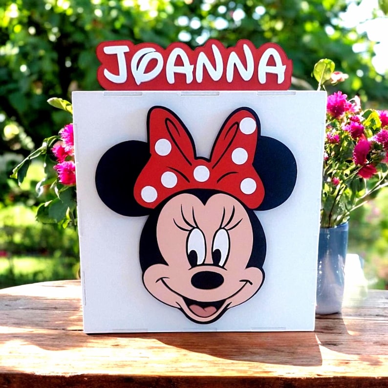 Op de afbeelding: Een witte doos met een Minnie Mouse-ontwerp, inclusief een rode strik met witte stippen. De naam "JOANNA" staat in witte letters op een rode achtergrond boven de Minnie Mouse-afbeelding. De doos staat op een houten oppervlak.