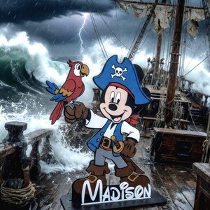 May include: A wooden cutout of Mickey Mouse dressed as a pirate, with a red and blue parrot perched on his hand. The pirate Mickey wears a blue hat with a skull and crossbones, a blue vest, and brown boots. The name "Madison" is displayed at the base.