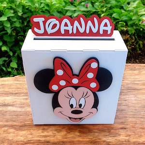 Op de afbeelding: Een witte houten doos met een gleuf aan de bovenkant, met een Minnie Mouse-ontwerp. De doos heeft een rood-wit naamplaatje met de tekst "JOANNA". Het Minnie Mouse-ontwerp bevat een rode strik met witte stippen.