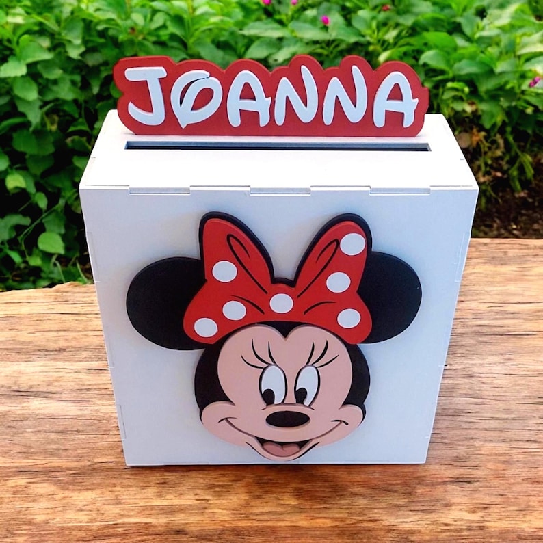 Op de afbeelding: Een witte houten doos met een gleuf aan de bovenkant, met een Minnie Mouse-ontwerp. De doos heeft een rood-wit naamplaatje met de tekst "JOANNA". Het Minnie Mouse-ontwerp bevat een rode strik met witte stippen.