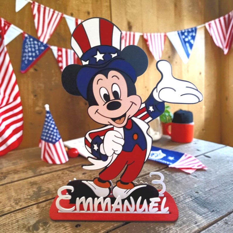 Puede incluir: Una figura patri&oacute;tica de Mickey Mouse con atuendo rojo, blanco y azul, incluyendo un sombrero de copa. La figura est&aacute; sobre una base roja con el nombre "Emmanuel" y el n&uacute;mero "3". Guirnaldas con la bandera americana decoran el fondo.