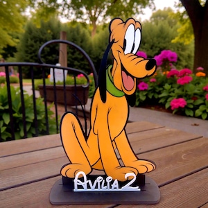 May include: A Pluto character cutout, featuring a smiling orange dog with black ears and a green collar. The cutout sits on a black base with the name "Avilia 2" in white. The background includes a wooden surface and a garden with pink flowers.