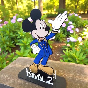 K&ouml;nnte beinhalten: Eine Mickey Mouse-Figur in blauem Anzug mit goldenen Schuhen und schwarzem Sockel. Auf dem Sockel stehen der Name "Kevin" und die Zahl "2" in wei&szlig;er Schrift. Der Hintergrund zeigt gr&uuml;nes Laub und Blumen.