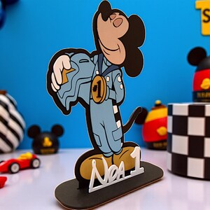 May include: A Mickey Mouse-themed cake topper. The figure is wearing a blue racing suit and a gold medal with the number 1. The base is black with the words "No. 1" in white. Other decorative items are visible in the background.