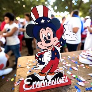 Puede incluir: Un recorte de Mickey Mouse con colores patri&oacute;ticos, con un dise&ntilde;o rojo, blanco y azul. El personaje lleva un sombrero de copa y saluda con la mano. El nombre "Emmanuel" se muestra en una base roja. Hay confeti esparcido alrededor de la base.