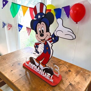 Puede incluir: Un recorte de madera de Mickey Mouse con colores patri&oacute;ticos, rojo, blanco y azul. El recorte est&aacute; sobre una base roja con el nombre "EMMANUEL" y el n&uacute;mero "3" en blanco. Un globo rojo y guirnaldas festivas en el fondo.