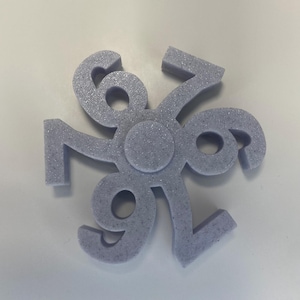 May include: A lavender-colored, three-dimensional decorative object with the number 696 arranged in a circular pattern. The object has a textured, speckled surface and a central circular design.
