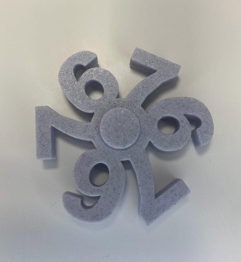 May include: A lavender-colored, three-dimensional decorative object with the number 696 arranged in a circular pattern. The object has a textured, speckled surface and a central circular design.