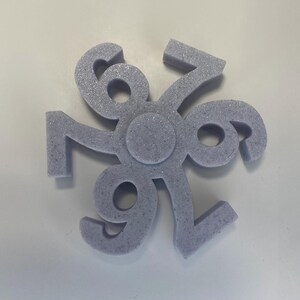 May include: A light purple, foam-like object with the numbers 6, 7, and 9 arranged in a circular pattern. The numbers are connected at a central point, creating a symmetrical design. The object has a textured surface and is set against a white background.