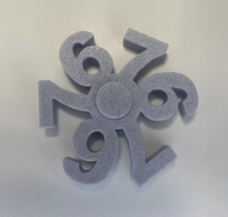 May include: A light purple, foam-like object with the numbers 6, 7, and 9 arranged in a circular pattern. The numbers are connected at a central point, creating a symmetrical design. The object has a textured surface and is set against a white background.
