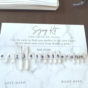 May include: A white sizing kit card for press-on nails with a marble pattern, featuring the text Sizing Kit FOR PRESS-ON NAILS. A strip of clear plastic press-on nails is attached, labeled with handwritten numbers from 0 to 14 for sizing.