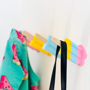 Könnte beinhalten: Drei popsicleförmige Haken an einer weißen Wand. Die Haken sind in Blau, Gelb und Pink mit einem Holzstiel lackiert. Ein blau-weiß gemustertes Hemd und ein schwarzer Riemen hängen an den Haken.