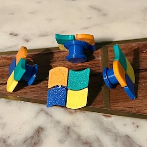 May include: A set of four colorful, plastic push pins arranged to resemble the Windows logo. Each pin has a blue base and a top section in shades of yellow, orange, green, and blue. The pins are on a wooden surface.