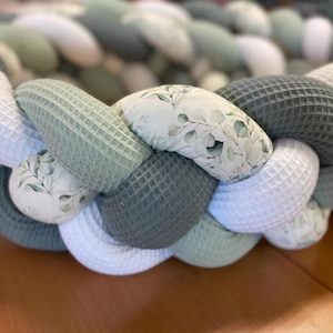 Könnte beinhalten: Ein geflochtenes Kissen mit einem weißen, grünen und grauen Muster. Das Kissen ist aus weichem Stoff und hat ein Blumenmuster.