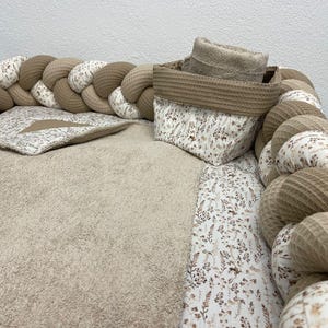 Könnte beinhalten: Ein geflochtener Babybett-Stoßfänger mit einem beige-weißen Blumenmuster. Der Stoßfänger ist aus weichem, plüschem Stoff gefertigt und schützt Babys perfekt vor Stößen und blauen Flecken. Der Stoßfänger ist außerdem mit einem kleinen, beigen Aufbewahrungskorb mit einem braunen Handtuch darin verziert.