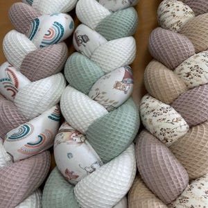 Peut inclure: Un oreiller noué tressé avec une variété de motifs de tissus, dont un motif arc-en-ciel, un motif d'animaux de la forêt et un motif floral. L'oreiller est fait d'un mélange de tissu gaufré et de tissu imprimé. L'oreiller est dans des tons de beige, marron, vert et blanc.