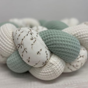 Könnte beinhalten: Geflochtener Baby-Bettumrandung in runder Form. Der Bettumrandung besteht aus Abschnitten aus weißem Waffelstoff, salbeigrünem Waffelstoff und Stoff mit einem botanischen Aufdruck von Zweigen und Blättern.