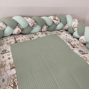 Può includere: Un nido per bambini con un paraurti intrecciato verde e bianco. Il paraurti è un mix di verde unito e tessuto fantasia con animali del bosco e alberi. Il nido comprende una coperta verde a trama waffle e un lenzuolo fantasia.