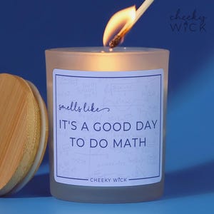 May include: A lit candle in a frosted glass jar with a wooden lid. The label reads "It's a good day to do math" with math equations in the background. A match is lighting the candle wick. The brand name "cheeky wick" is visible.