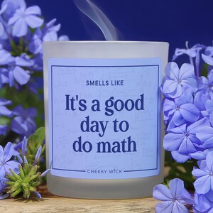 May include: A frosted glass candle with a light blue label featuring the text "SMELLS LIKE It's a good day to do math" and "CHEEKY WICK" in dark blue font, with faint mathematical equations in the background and a thin wisp of smoke rising.