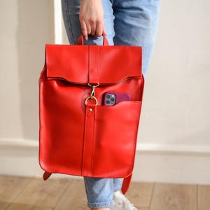 Może przedstawiać: Jasnoczerwony skórzany plecak z klapką i przednią kieszenią. Torba ma złote zapięcie, a w kieszeni widoczny jest bordowy telefon. Torbę trzyma osoba ubrana w niebieskie dżinsy i białe trampki.