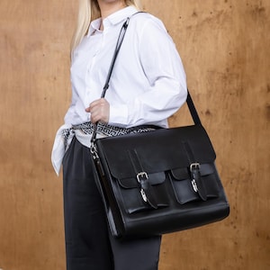 May include: A black leather satchel bag with three silver buckles. The bag has a long shoulder strap and is being carried by a person wearing a white button-down shirt and black pants. The background is a light brown wood panel.