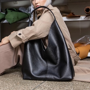 Puede incluir: Un bolso tote grande de cuero negro con un asa curva. El bolso está sobre un suelo de hormigón. Estanterías en el fondo están llenas de rollos de tela de varios colores, incluyendo rojo, marrón y verde.