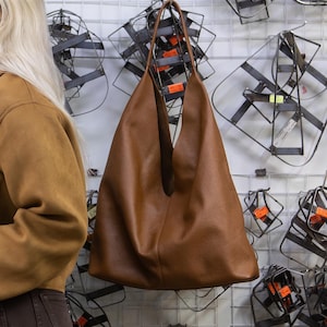 Könnte beinhalten: Eine Frau trägt eine braune Wildlederjacke, eine braune Hose und eine Sonnenbrille. Eine große braune Ledertasche hängt an der Wand. Der Hintergrund zeigt Metallobjekte und ein Gitter.