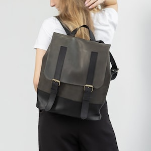Puede incluir: Una mochila de cuero verde oscuro y negro con correas ajustables y hebillas doradas. La mochila tiene un cierre de solapa y un asa superior. La bolsa se lleva sobre los hombros.