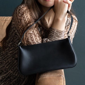 May include: A woman wearing a brown and white patterned blouse sits on a brown sofa. She is holding a black leather handbag with a thin strap.