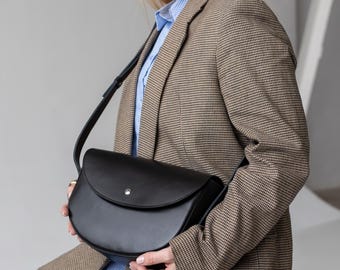 Puede incluir: Un bolso bandolera de cuero negro con una silueta curva en forma de silla de montar y un cierre de presión plateado en la solapa frontal. El bolso cuenta con una correa ajustable y un acabado liso y mate, con un diseño sencillo y elegante.